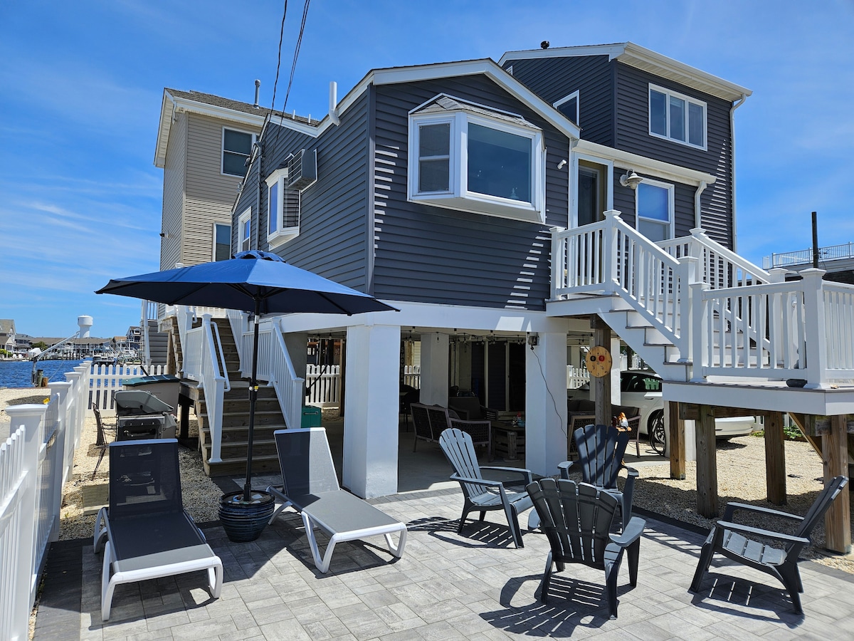 The outdoor space features a paved patio area with several lounge chairs positioned for relaxation. An umbrella provides shade over a seating area, while the building's elevated structure offers views of the surrounding area and water.