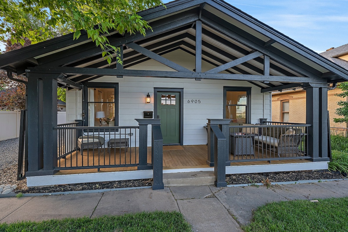 The bungalow showcases a covered front porch, featuring seating arrangements that invite relaxation. The home's exterior is highlighted by a mix of dark beams and light clapboard siding, with a charming front door centered between large windows that allow natural light to fill the interior.
