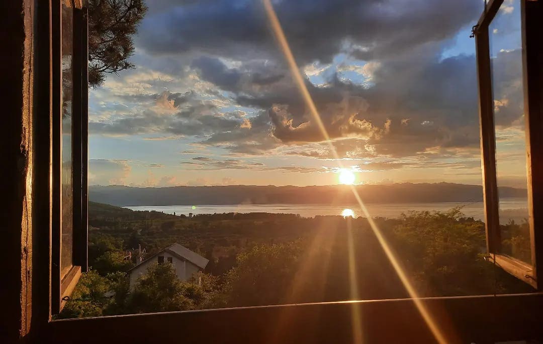 A view from an open window captures a stunning sunset over the tranquil Lake Ohrid, accented by gentle cloud formations and golden rays of sunlight. Lush greenery and distant hills are visible, providing a beautiful natural backdrop.