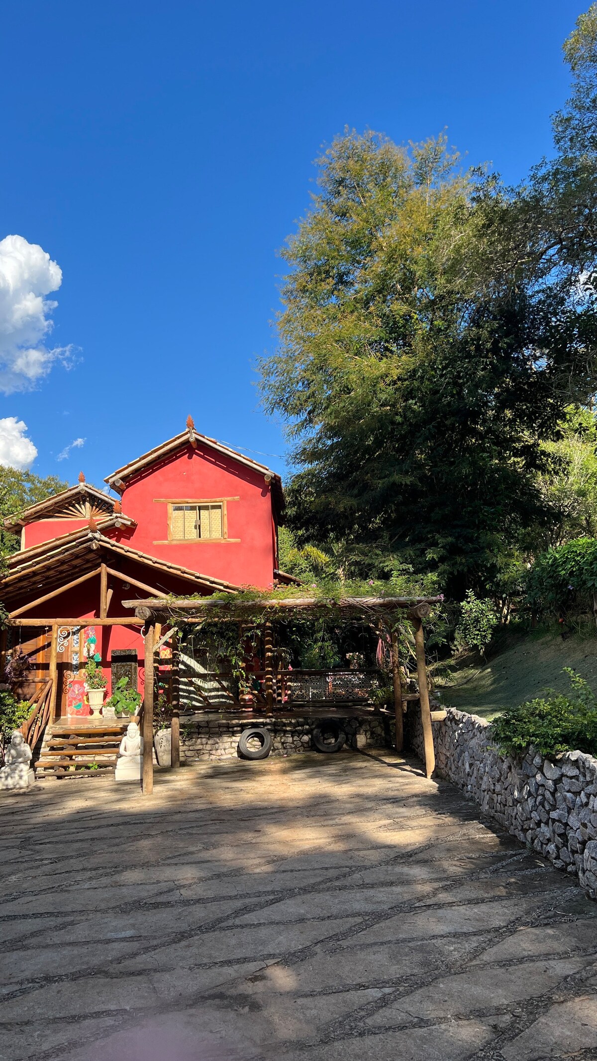 A charming rustic building features a red facade with wooden accents, surrounded by lush greenery. A pergola extends over the entrance, providing shade. The landscape includes a stone wall and trees, with a clear blue sky visible above.