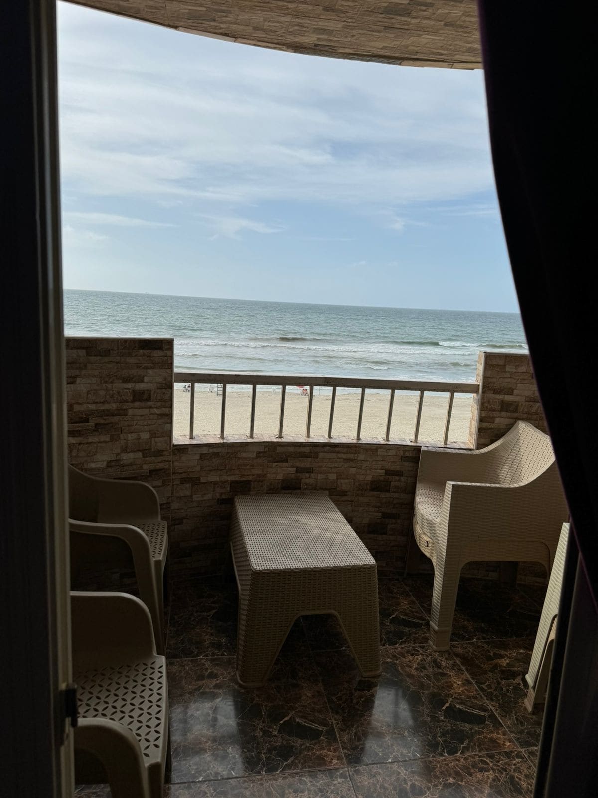 A spacious balcony features a view of the ocean and beach, with several lightweight chairs and a low table positioned for relaxation. The marble flooring adds elegance, while ocean waves can be seen gently lapping at the shore in the distance.