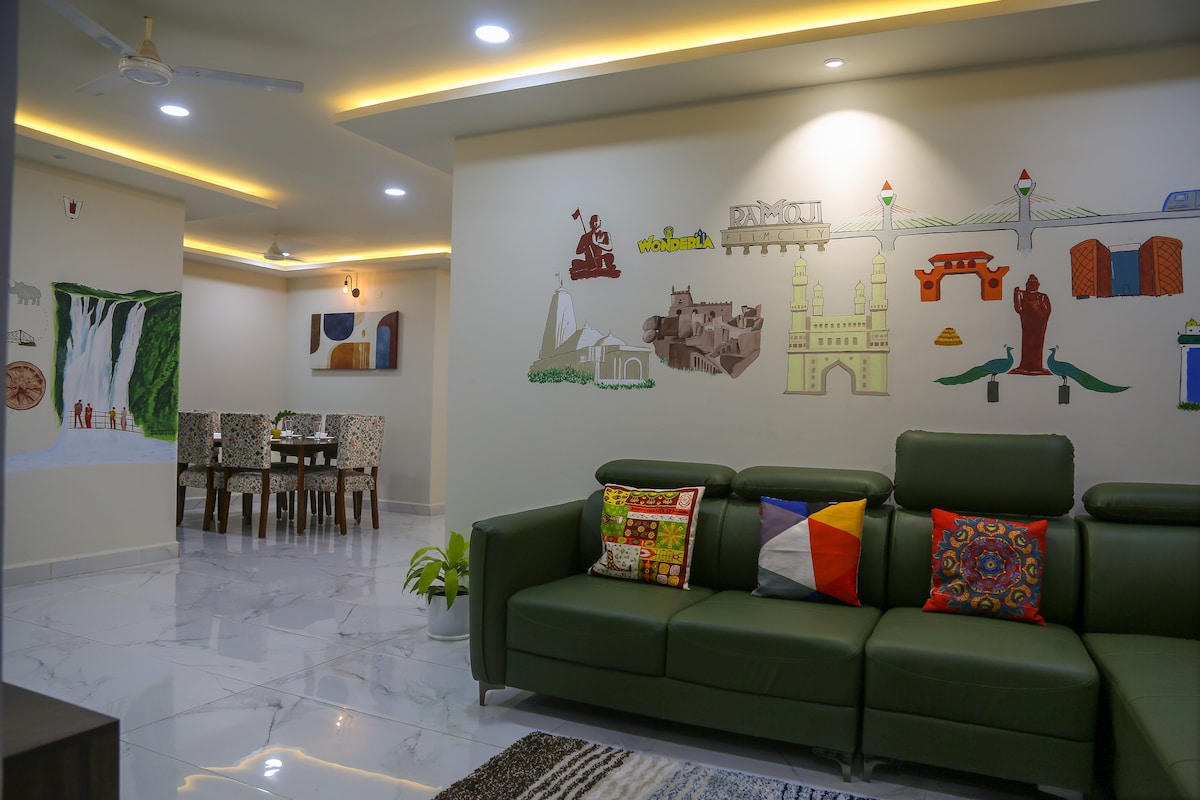 A modern living area is displayed, featuring a green sofa adorned with colorful cushions. A mural representing various landmarks and cultural symbols is positioned on the wall, providing a vibrant backdrop. A dining area with a table and patterned chairs is visible in the distance.
