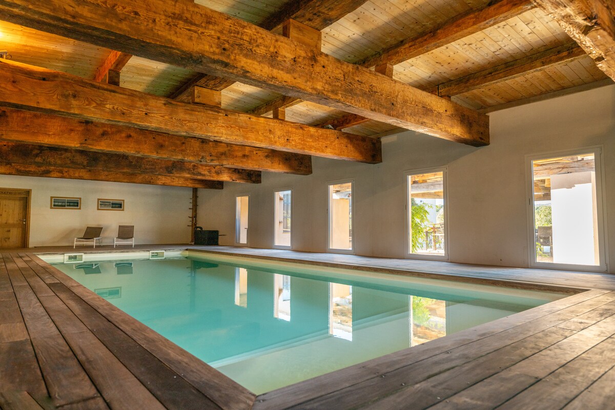 An indoor heated swimming pool is framed by wooden beams and features large windows allowing natural light to flood the space. A wooden deck surrounds the pool, and two lounge chairs are positioned nearby, offering a relaxed atmosphere.