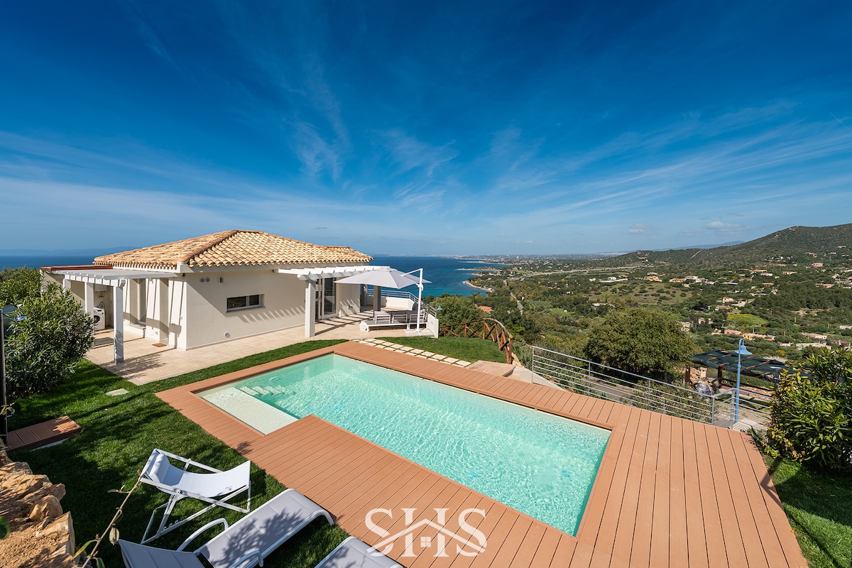A private villa with a tiled roof overlooks a turquoise swimming pool and surrounding greenery. A spacious deck frames the pool, while the scenic coastline and hills are visible in the background under a clear blue sky.