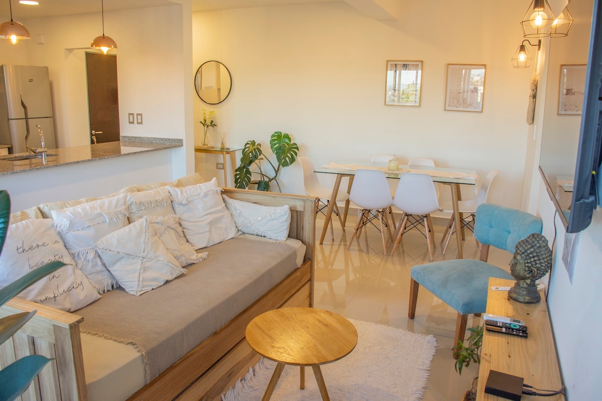 A bright living area features a comfortable sofa adorned with various cushions, complemented by a wooden coffee table. A stylish dining table with white chairs is visible in the background, along with decorative wall art and plants, creating a welcoming ambiance.