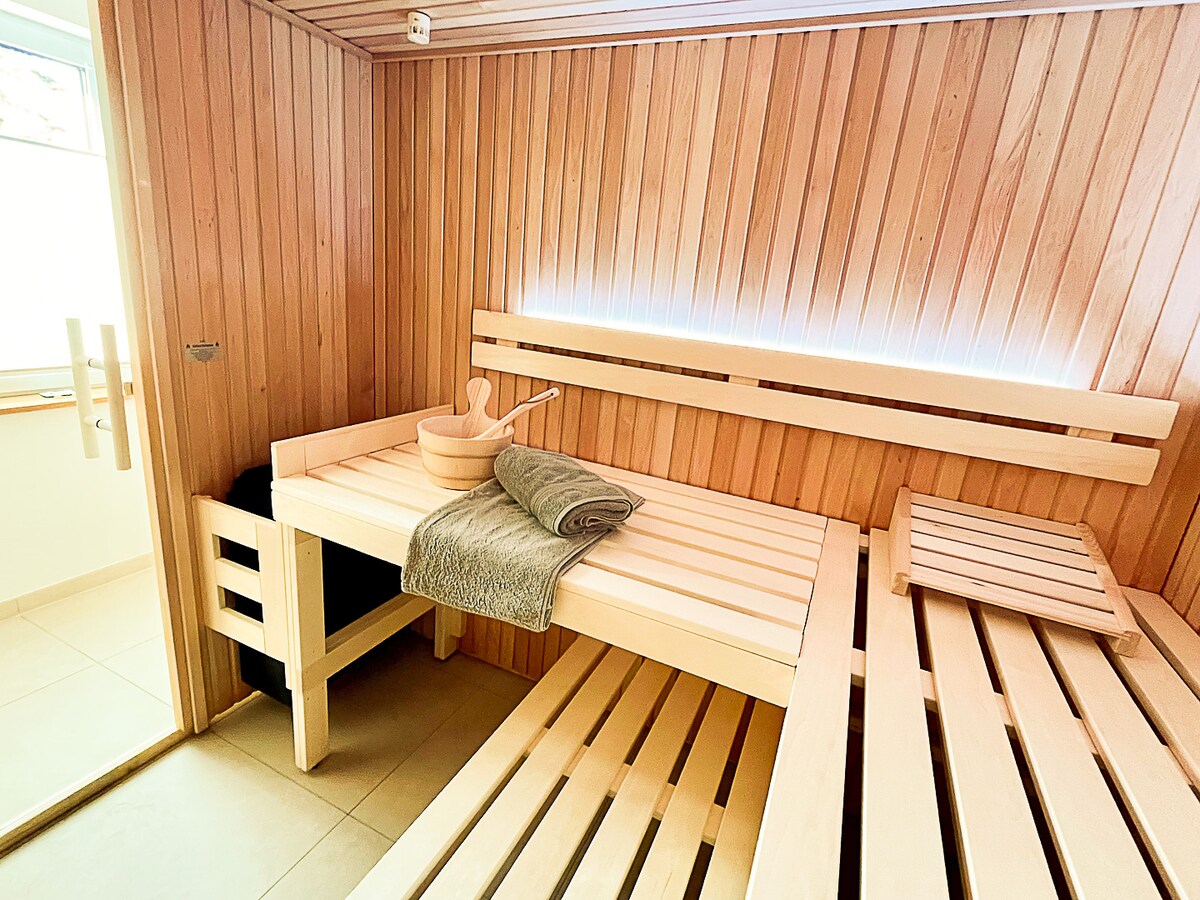 A wooden sauna is featured, complete with multiple benches arranged for relaxation. A bucket and a rolled towel are positioned on one of the benches. Soft lighting enhances the warm wood tones, creating an inviting space for unwinding.