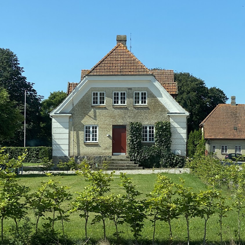 The charming historic house features a gabled roof and a welcoming red door. Lush greenery frames the entrance, while well-maintained landscaping and nearby ancient trees provide a serene backdrop. The facade showcases multiple windows, reflecting natural light in the peaceful surroundings.