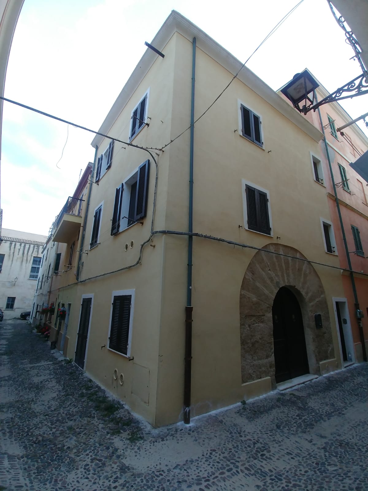 A three-story building is shown from an angle, highlighting its warm yellow exterior and traditional dark shuttered windows. An arched stone doorway is visible, framed by light stonework. The cobblestone street adds texture to the environment, while overhead electrical lines create a sense of urban living.