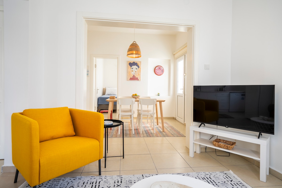 A welcoming living area features a bright yellow armchair paired with a small black side table. A modern TV is positioned on a white console. In the background, a dining area with a light wood table and four chairs is partially visible, accentuated by soft lighting and colorful artwork.