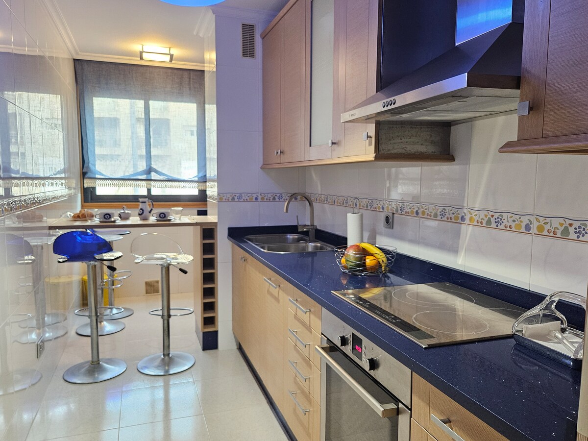 A modern kitchen is featured, equipped with wooden cabinetry and sleek blue countertops. Stainless steel appliances, including a stovetop and oven, are visible. A breakfast bar with high stools offers additional seating, and natural light enters through large windows with sheer coverings.