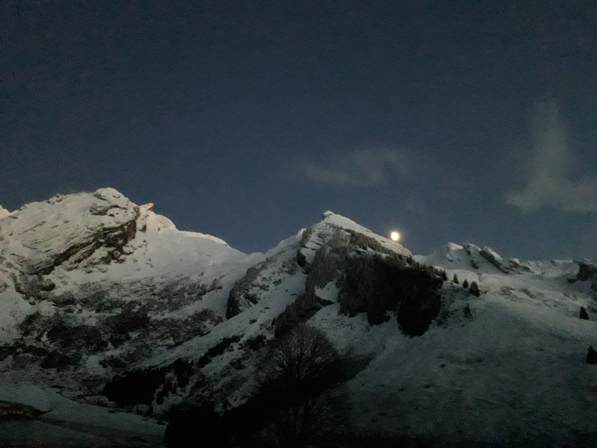 Snow-covered mountains rise sharply against a night sky, where a full moon casts a soft glow over the landscape. The rugged peaks and gentle slopes create a serene atmosphere, enhancing the calmness of the scene.