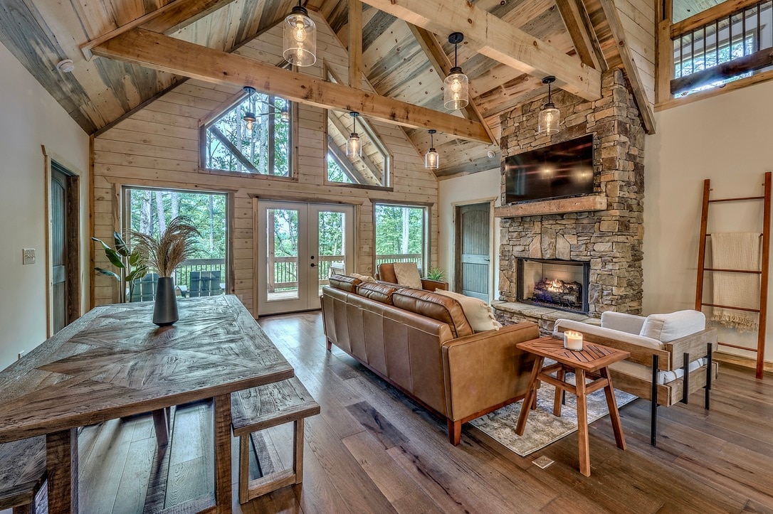 A spacious living area features large windows that provide natural light and views of the surrounding trees. A stone fireplace anchors the room, complemented by rustic wooden beams and modern furnishings, including a comfortable sofa and a wooden dining table.