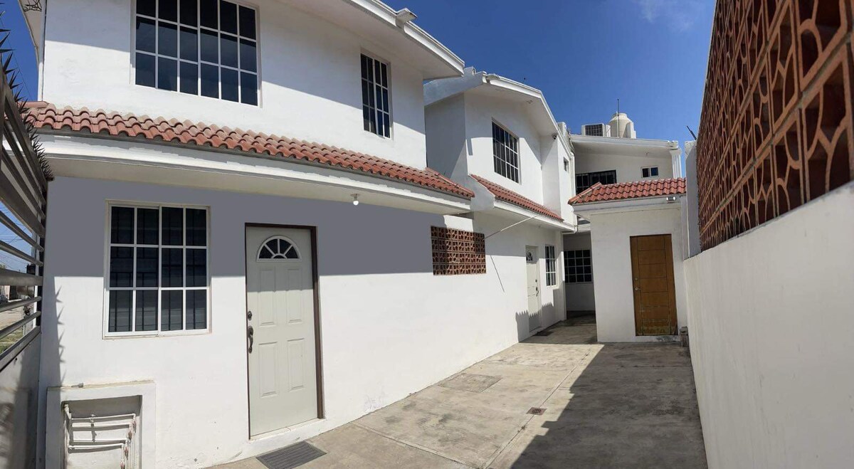 A well-maintained property is depicted, featuring a white exterior with multiple windows allowing natural light. A paved courtyard can be seen, with a door leading into the house and another door that is partially visible. The roof is adorned with red tiles.