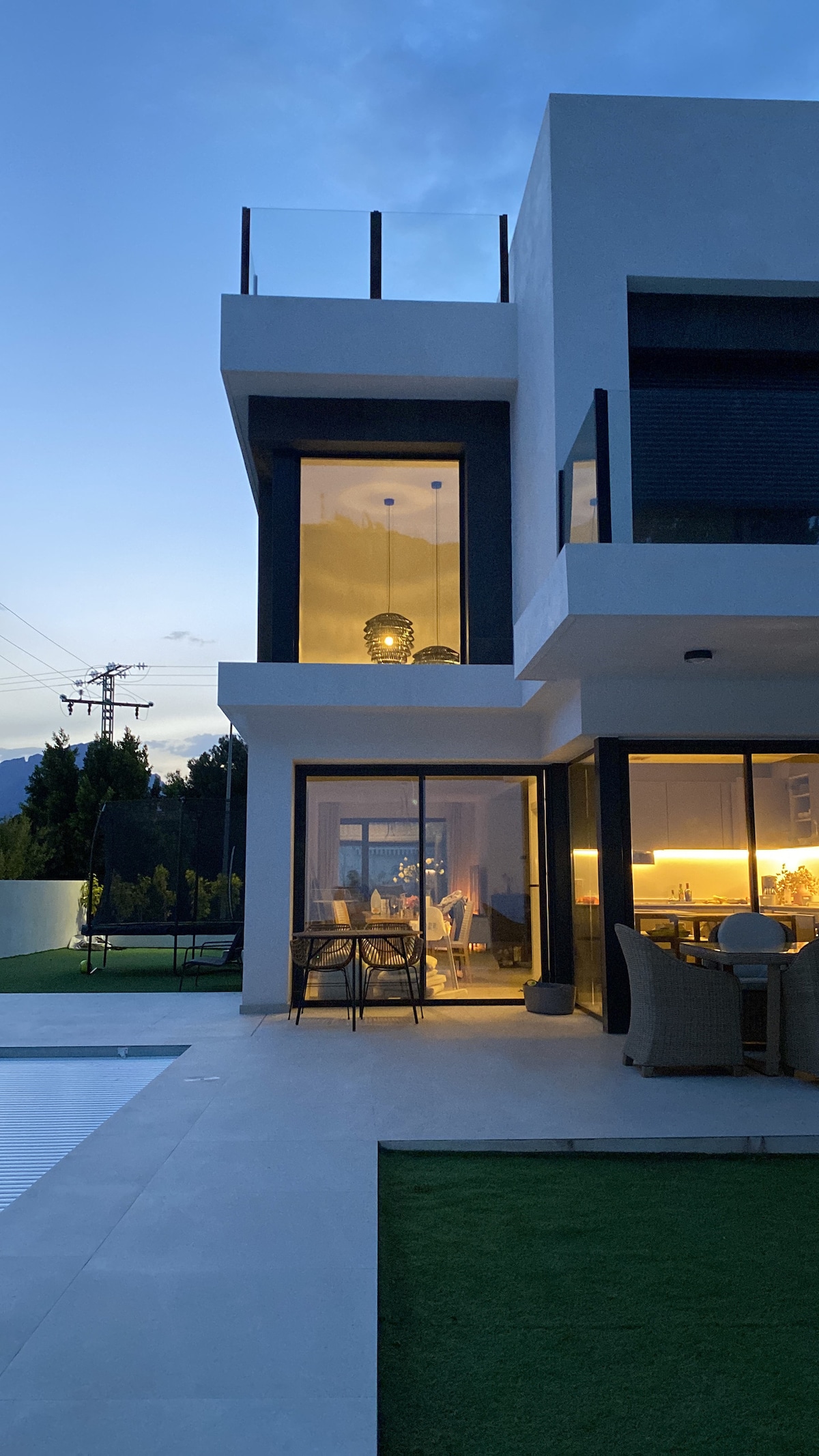 The exterior of a modern house is shown at dusk, featuring large glass walls that reveal warm indoor lighting. A spacious terrace can be seen above, while the pool and garden area are visible in the foreground, emphasizing a peaceful outdoor environment.