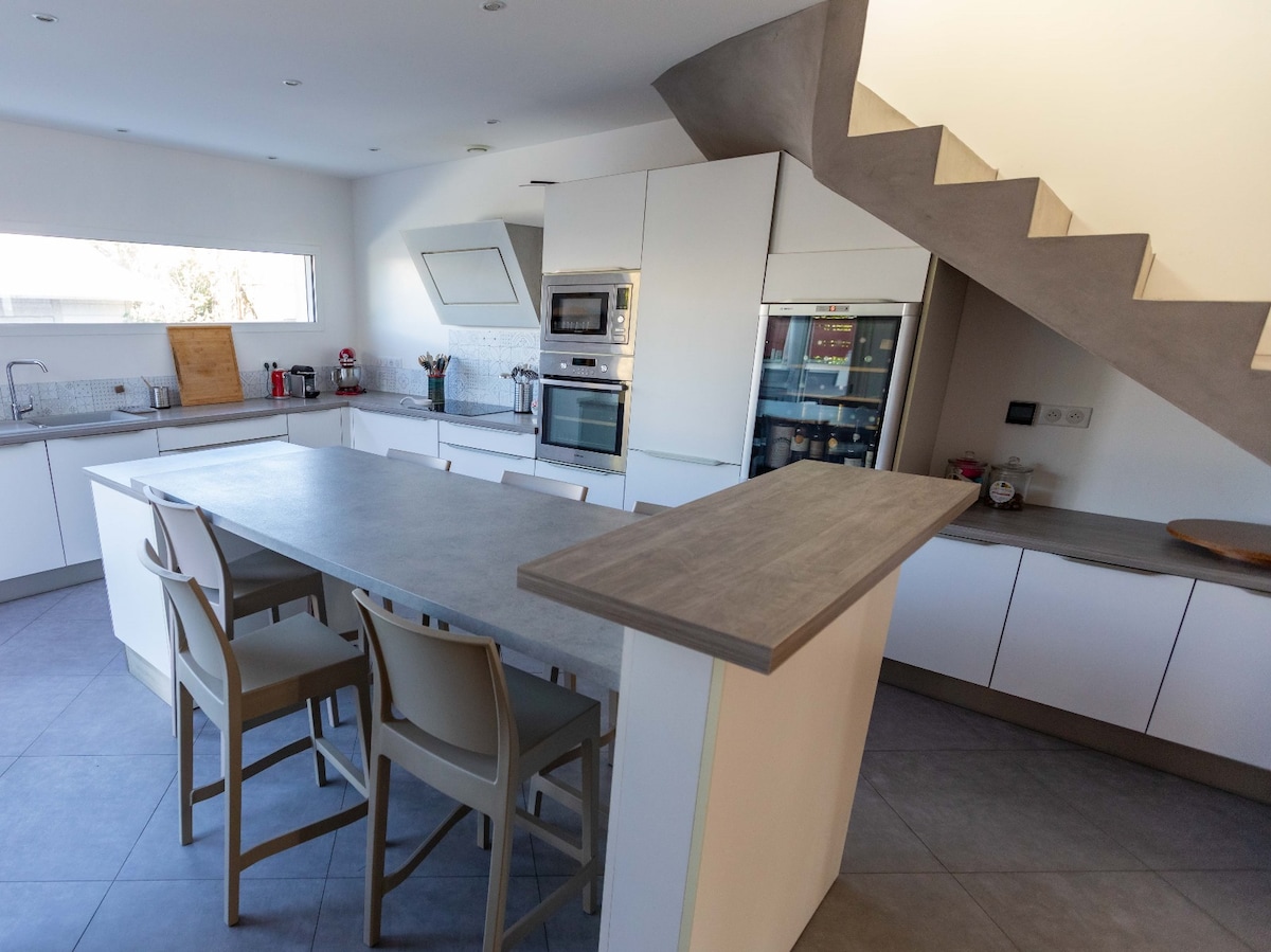 A modern kitchen features a spacious island counter with seating for four. Sleek cabinetry and stainless steel appliances are complemented by ample natural light from the large window. A staircase is visible, leading to the upper level.