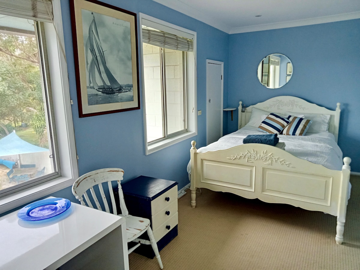 A spacious light-filled room features a large queen size bed with decorative pillows. A desk, white chair, and a small nightstand are provided. Two windows with blinds offer views of greenery outside, while a round mirror adorns the wall above the bed.