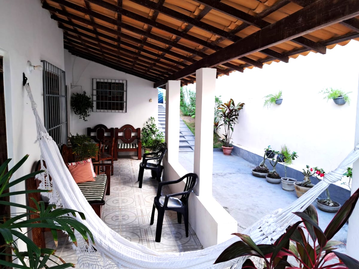 A shaded terrace features a hammock and seating arrangements, including chairs and a wooden table. Potted plants add greenery to the space. The tiled flooring and textured ceiling create a warm atmosphere, while a staircase leads to an outdoor area beyond.