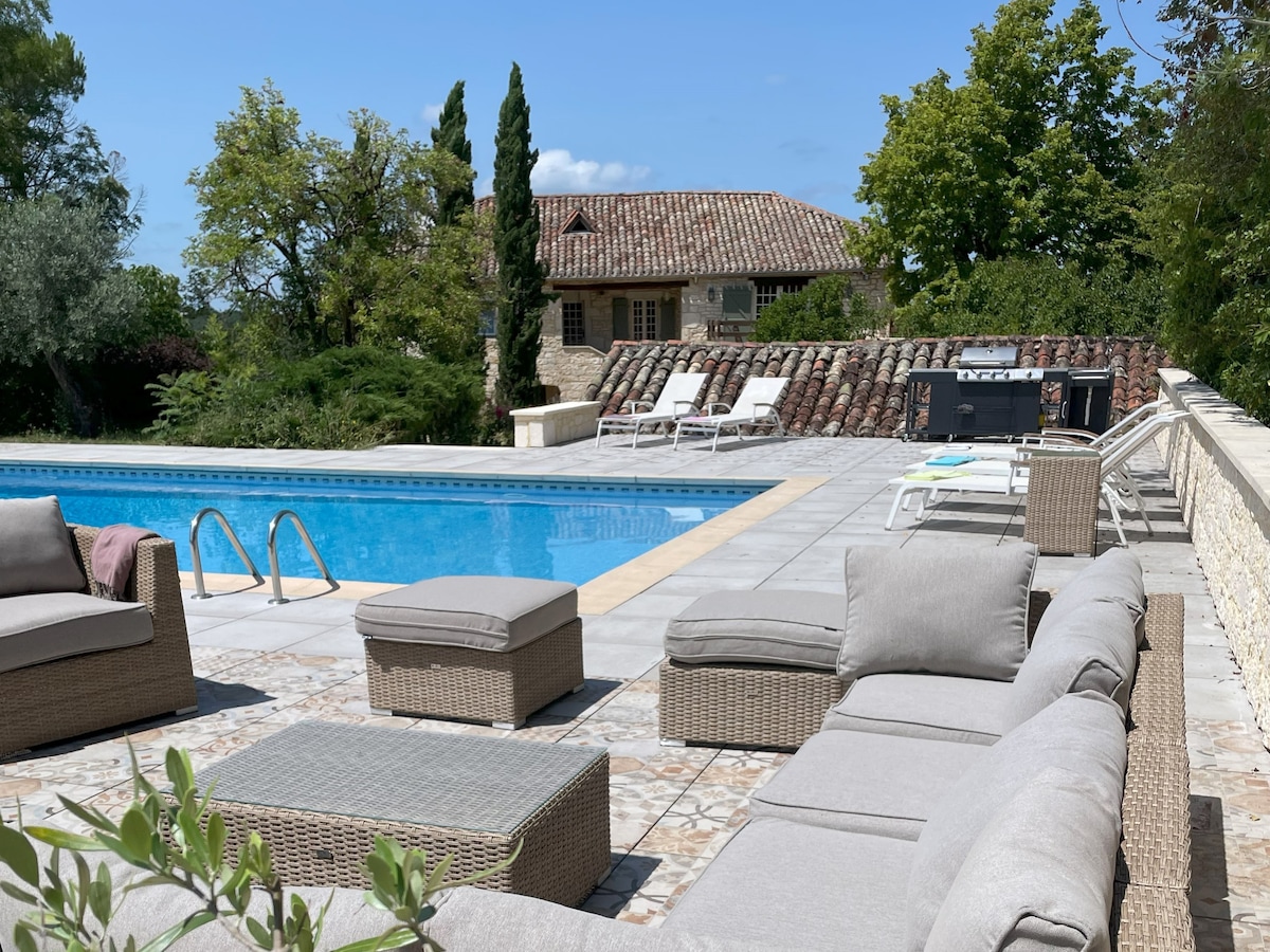 A spacious outdoor area is shown with a heated pool, surrounded by sun loungers and modern seating. A barbecue station and greenery are visible, creating an inviting space for relaxation and enjoyment.