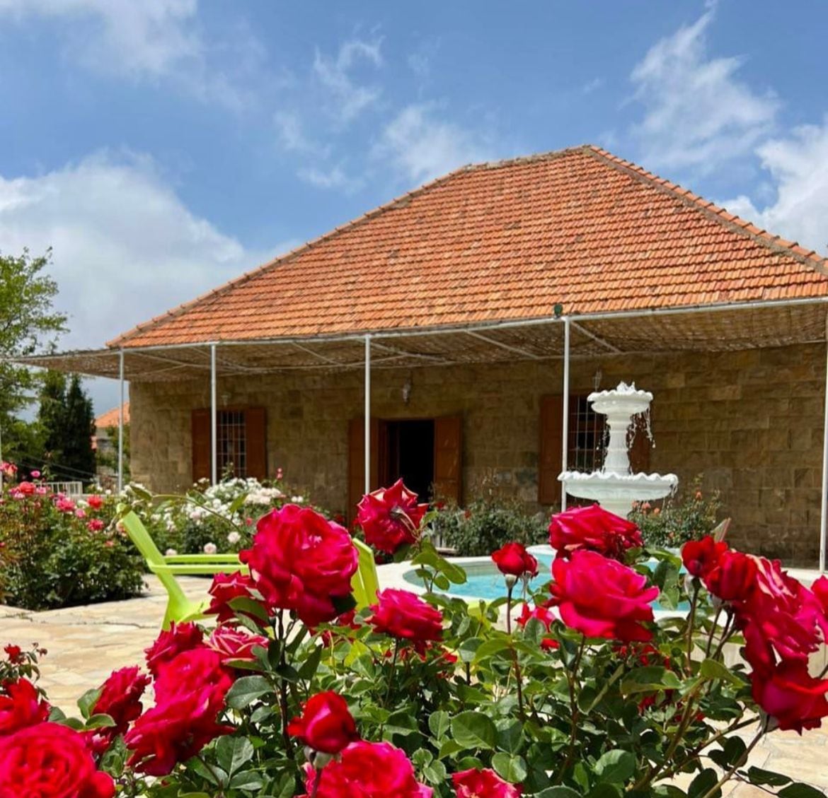 A traditional Lebanese home is framed by vibrant red roses in full bloom. The stone exterior and red-tiled roof complement the lush garden. A fountain can be seen in the outdoor area, surrounded by greenery and inviting seating.