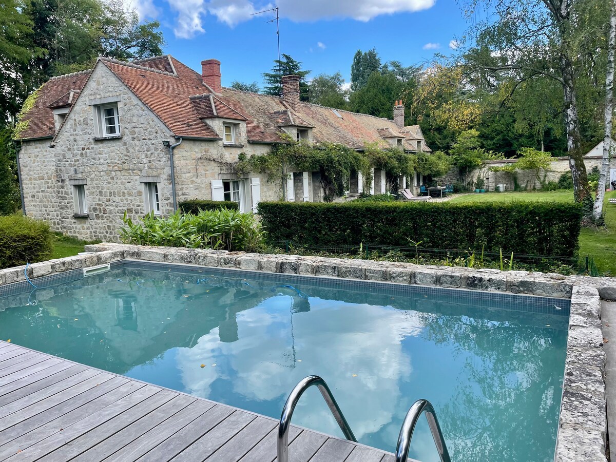 A stone house with a sloping roof is visible in the background, surrounded by lush greenery. A serene pool in the foreground reflects the blue sky, with steps leading into the water. The garden features well-maintained hedges and a variety of trees, adding to the tranquil atmosphere.