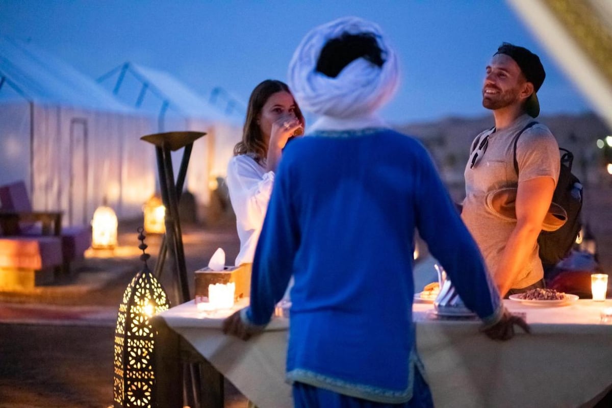 An evening scene is captured at the desert camp, where guests are engaged in conversation by a beautifully set table. Soft candlelight glows from ornate lanterns, creating a warm atmosphere. Tents are visible in the background, adding to the immersive desert experience.