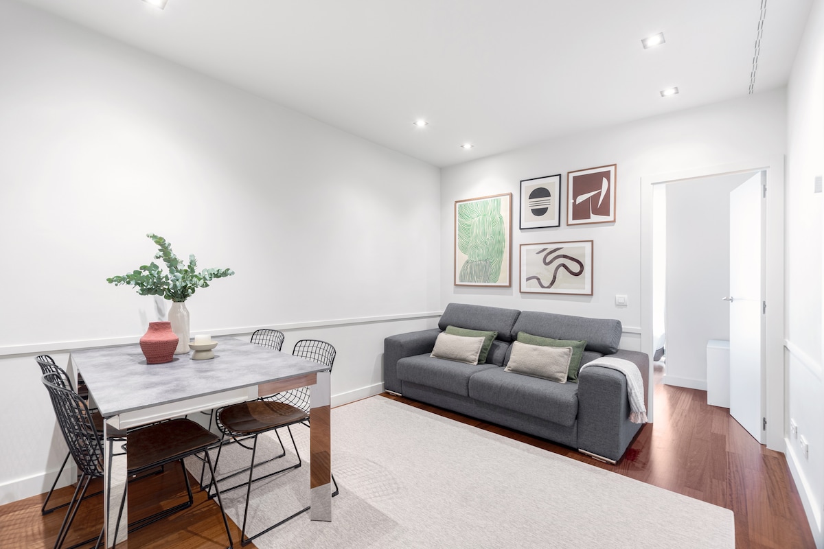 A modern living and dining area features a sleek dining table surrounded by black wire chairs. A comfortable gray sofa is adorned with cushions, positioned against a wall displaying framed art pieces. Light-colored walls enhance the bright feel of the space.