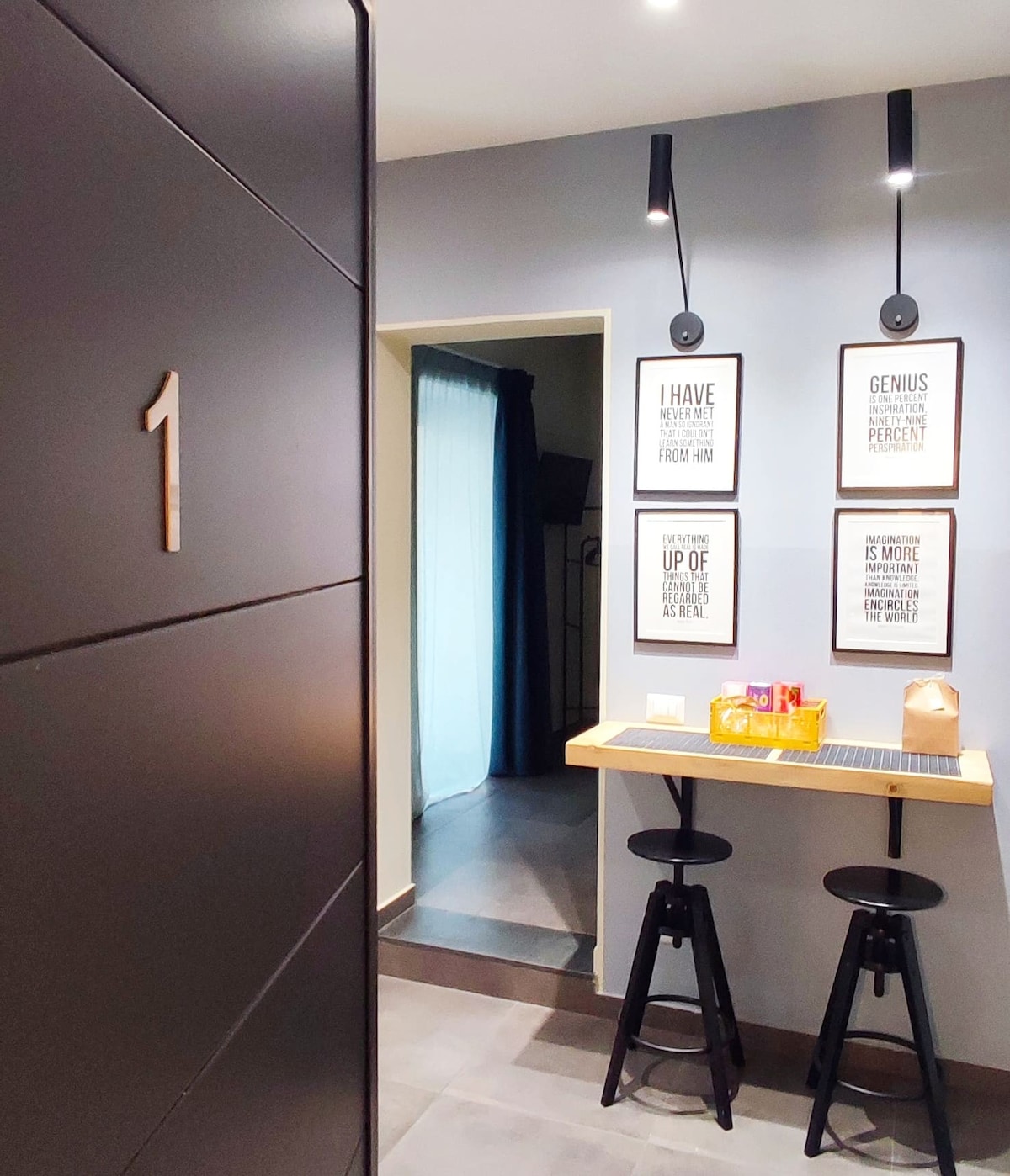 The entrance area features a small kitchen counter with two black stools, complemented by decorative wall art. A doorway leads to another room, while a dark cabinet bears the number '1' prominently. Natural light enters through a window partially covered by a curtain.