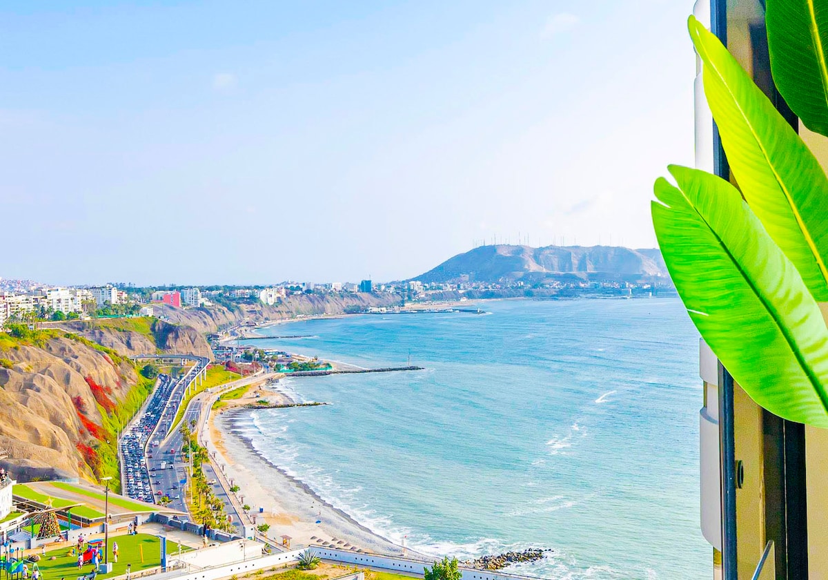 A sweeping view of the Pacific Ocean is visible from the balcony, showcasing the coastline with gentle waves and sandy beaches. The lush greenery in the foreground adds a touch of nature, while distant hills create a picturesque background.