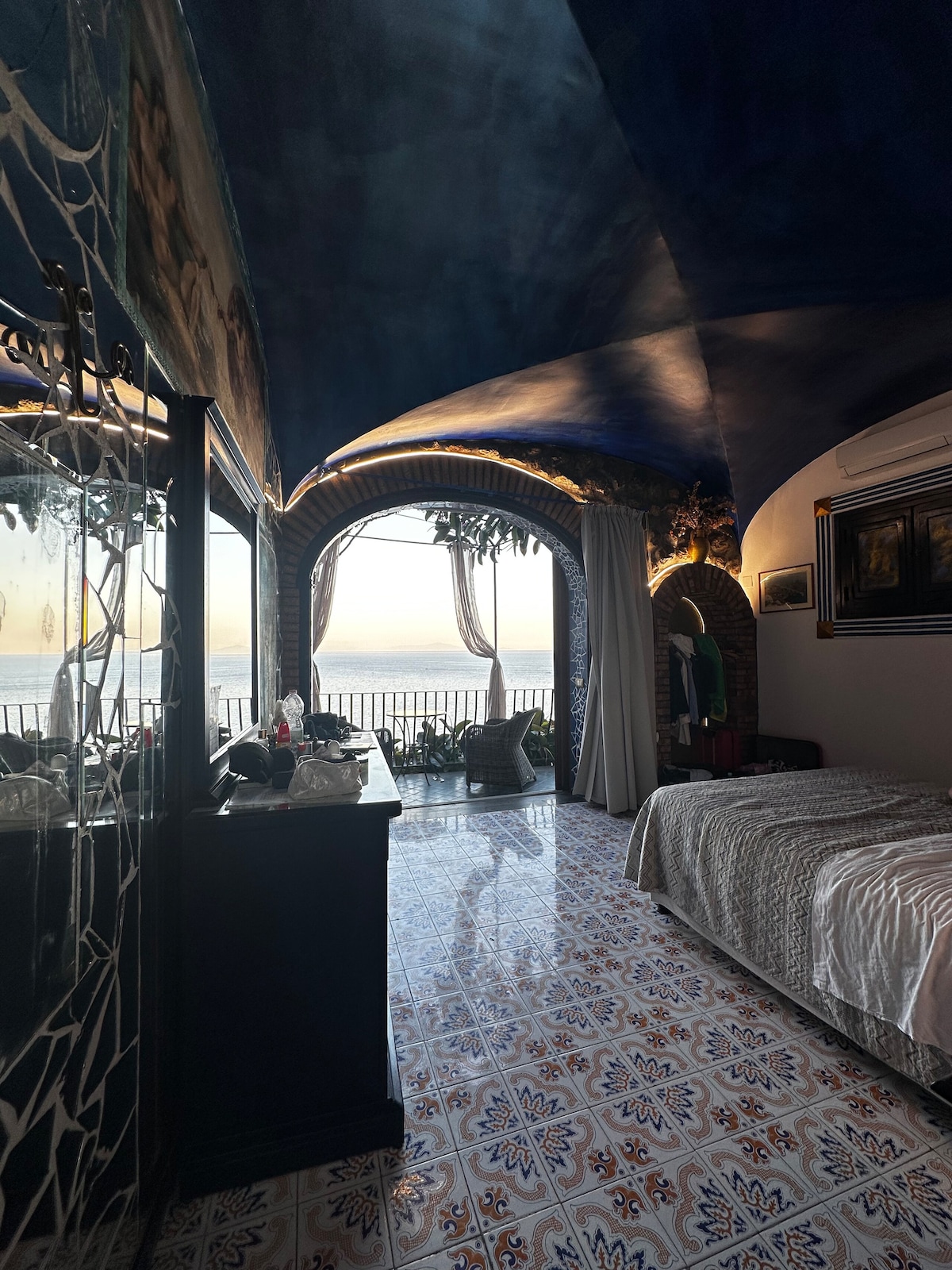 A spacious room features a high, arched ceiling with soft blue hues, creating an airy ambiance. Large windows offer views of the sea, while vibrant ceramic tiles adorn the floor, contributing to the local style. A dark wood dresser complements the decor.