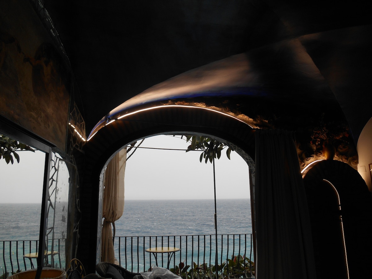 The image captures a cozy interior space with arched ceilings, where soft lighting accents the contours. Large windows frame a stunning view of the sea, with a balcony visible, offering a glimpse of serene waters. Delicate curtains sway gently, enhancing the tranquil ambiance.
