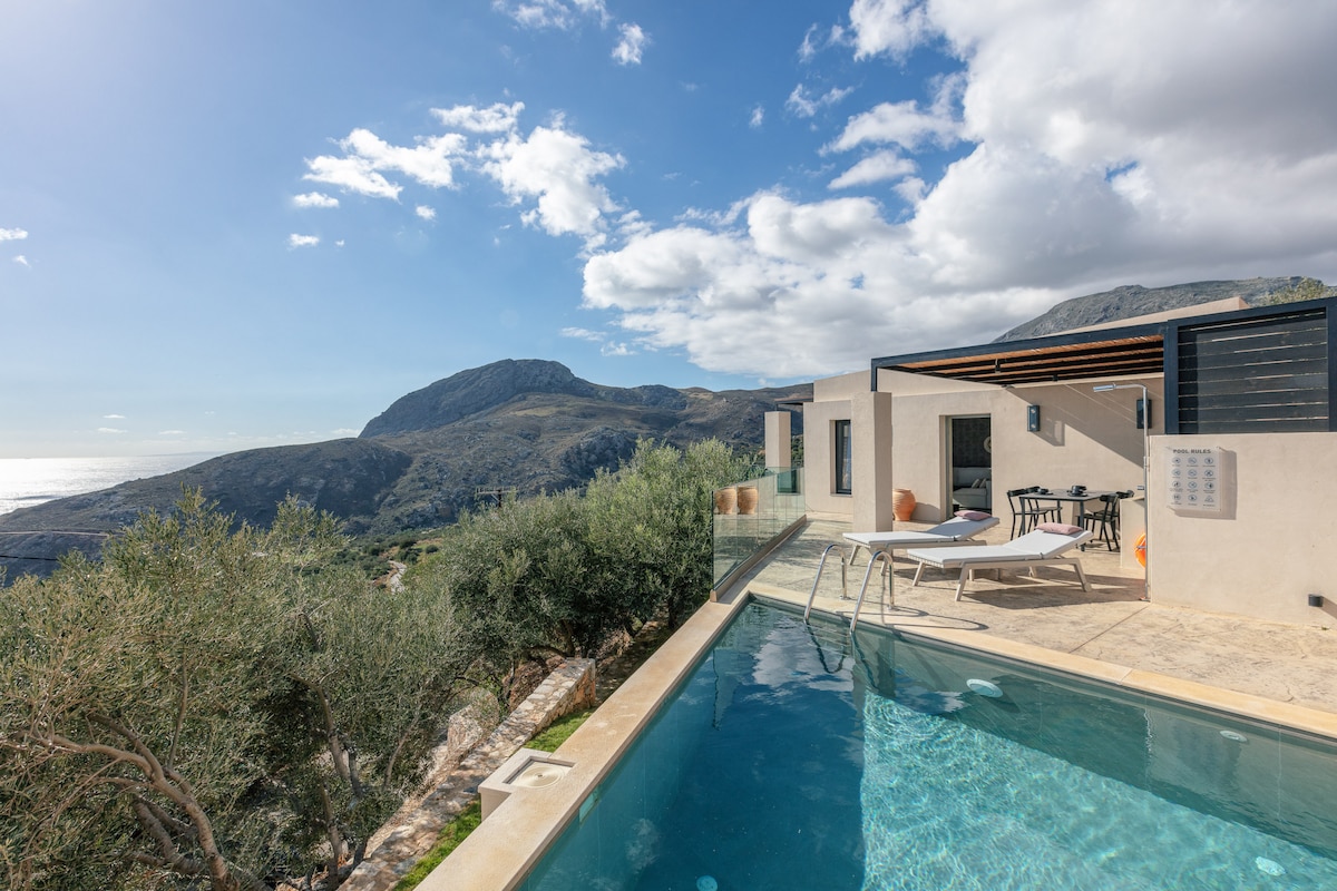 A private swimming pool is set against a scenic background of rolling hills and the sea. Modern sun loungers are placed beside the pool, while a contemporary villa is visible in the distance, featuring large glass doors that invite natural light.