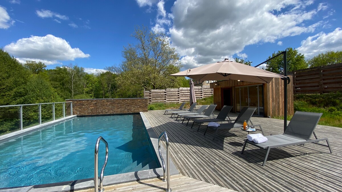 An inviting outdoor wellness area features a heated swimming pool surrounded by wooden decking and six lounge chairs. A shaded area with a canopy provides shelter alongside the pool, while lush greenery is visible in the background under a partly cloudy sky.