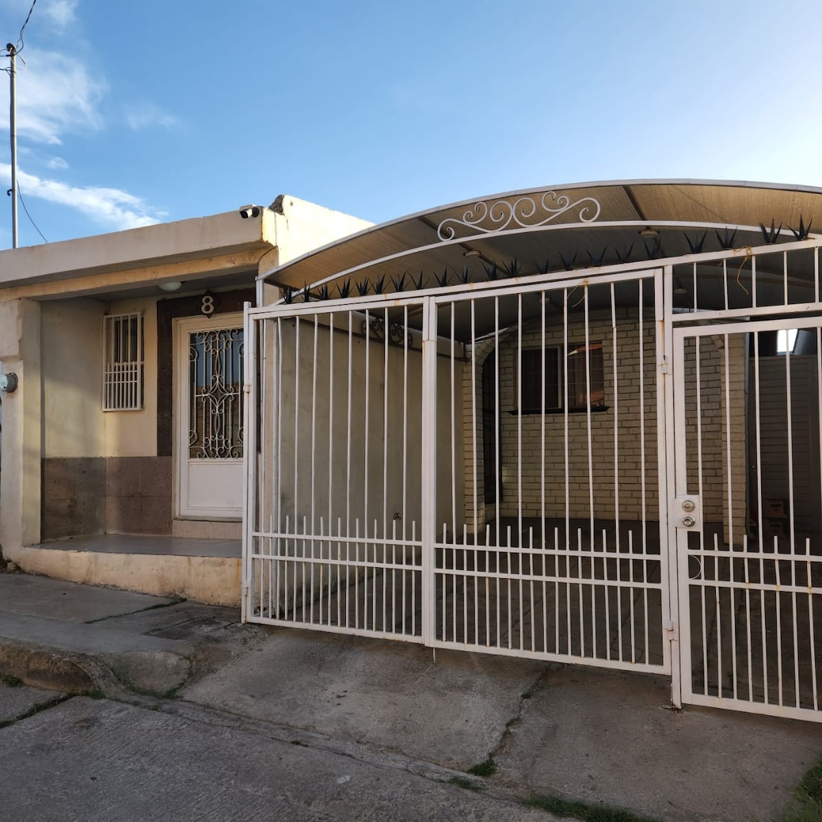 A single-story home is enclosed by a white wrought iron gate, providing privacy. The entrance features a decorative front door, while the driveway is covered by a canopy. The exterior showcases a simple design with a light-colored finish, and the surrounding area features a clear blue sky.