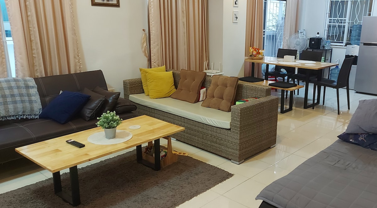 The living area features a mix of seating options, including a dark sofa and a light, cushioned wicker couch adorned with colorful pillows. A central wooden coffee table sits atop a soft rug, while a dining table is visible in the background.
