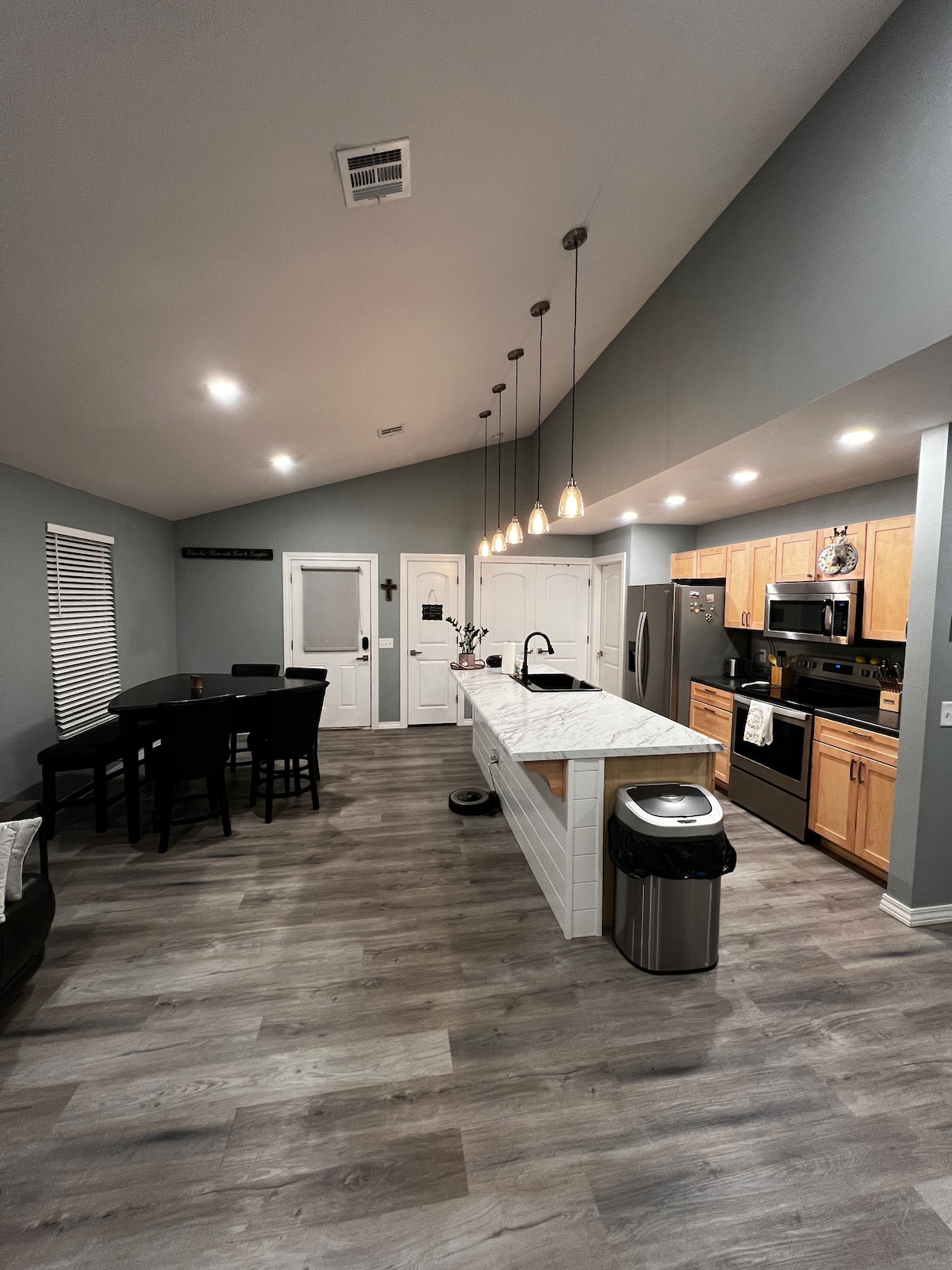 An inviting kitchen and dining area showcases modern fixtures and finishes. The kitchen features a spacious island with stools, stainless steel appliances, and pendant lighting. A dining table accommodates six, and soft lighting enhances the open layout, creating a functional space for gatherings.