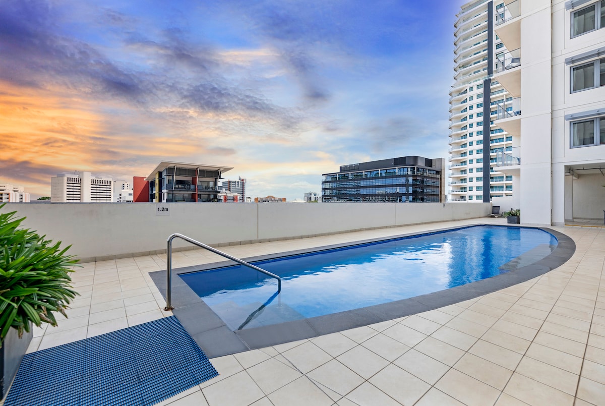 An outdoor swimming pool glistens under a colorful sunset, framed by modern high-rise buildings. The inviting water is accessible via a stainless steel railing, set on a spacious, tiled deck surrounded by lush greenery.
