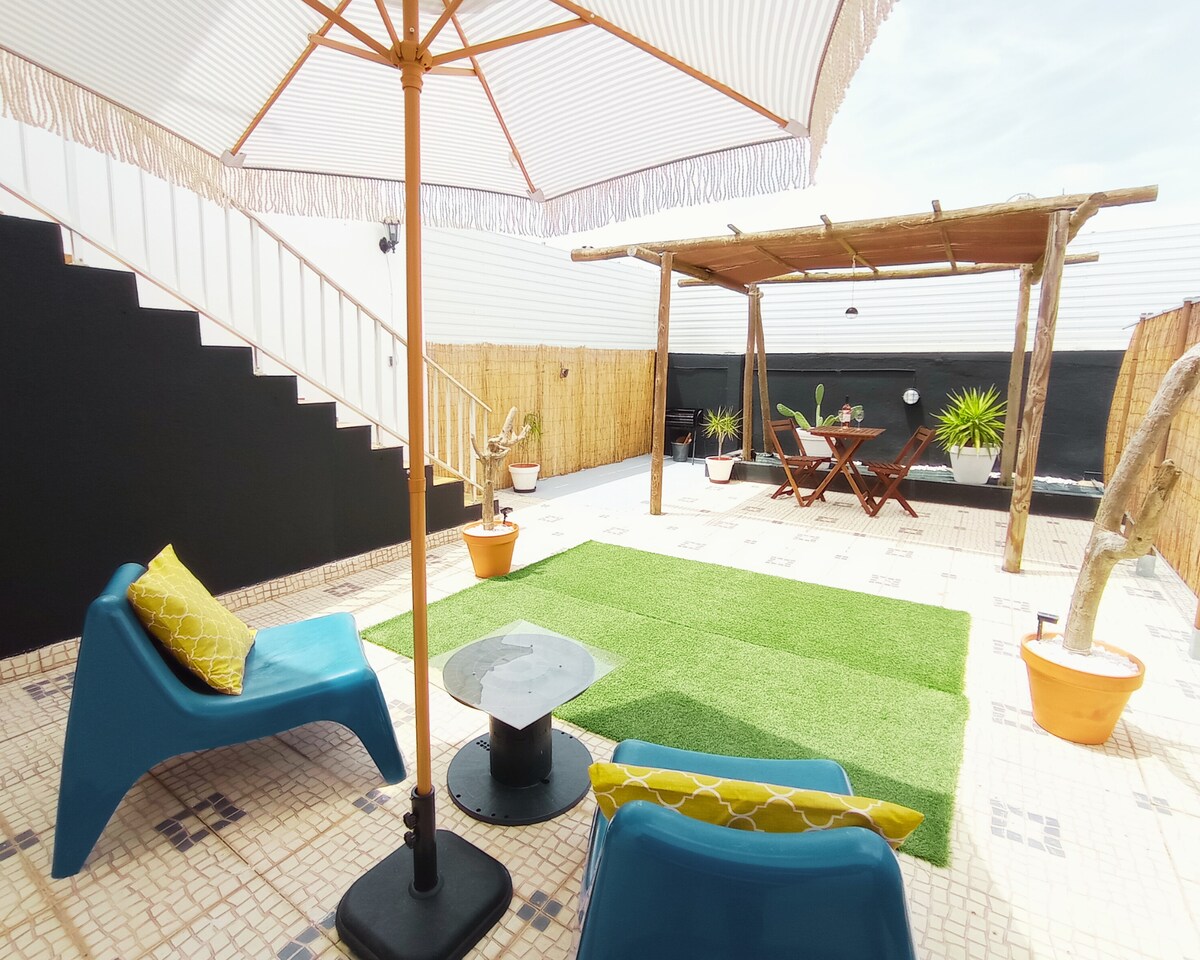 An outdoor lounge area is depicted with modern seating in vibrant blue surrounding a small table. A large umbrella provides shade over the seating. A thatched roof structure shelters a dining table with chairs, while potted plants add greenery against the backdrop of a black wall.