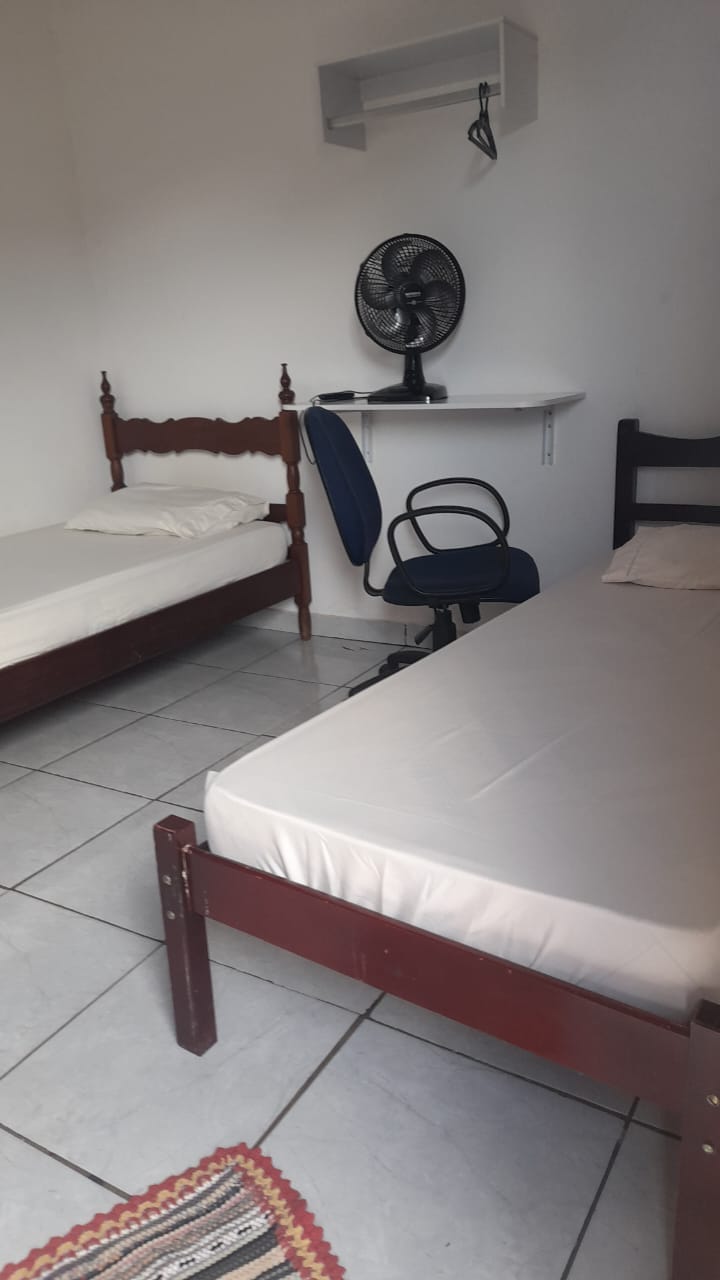 The room contains two individual beds with simple wooden frames, positioned slightly apart. A small desk is equipped with a chair, and a standing fan is visible nearby. A tiled floor and a neutral wall color contribute to the room's minimalist design.
