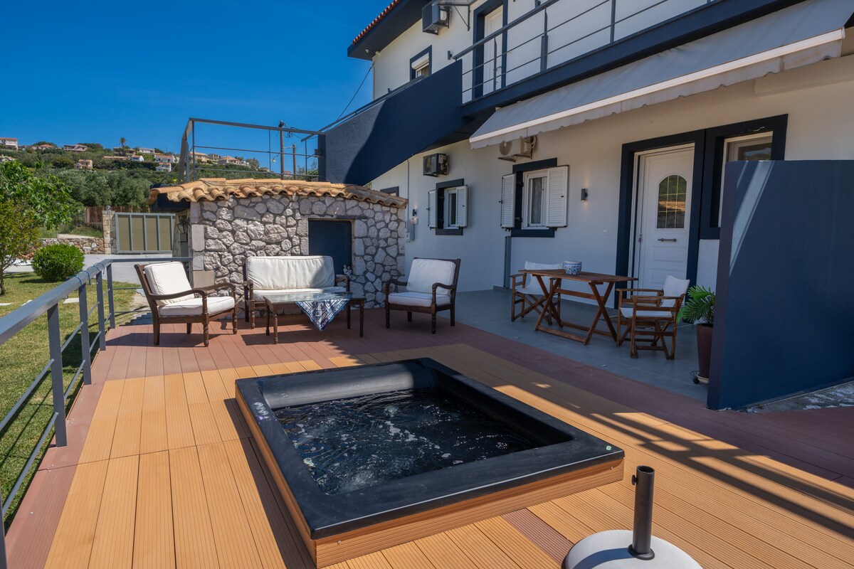 A private terrace features a hot tub, surrounded by outdoor seating and a small dining set. The area is enhanced by a stone barbecue, and large windows provide ample light. The surrounding greenery and distant hills contribute to a serene outdoor setting.