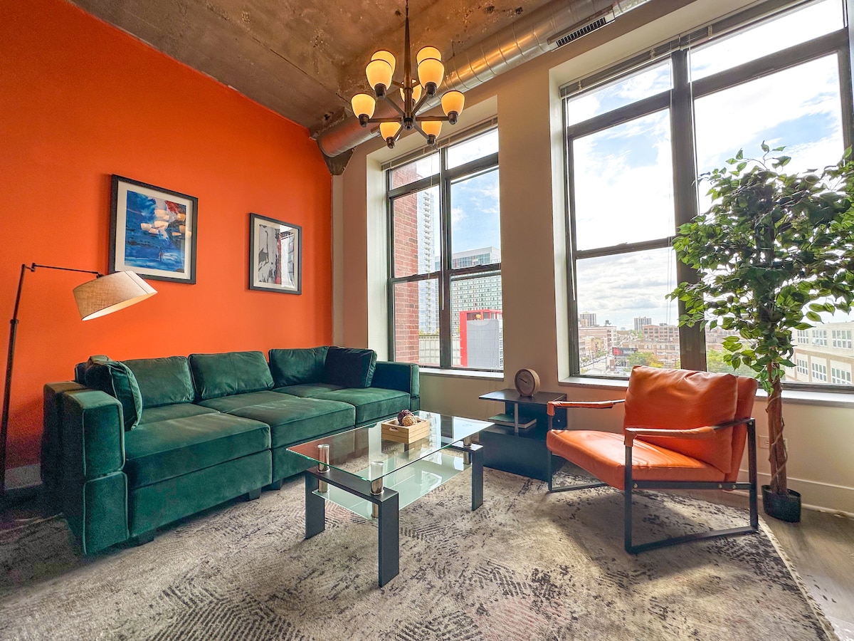 A modern living area features a plush green sectional couch, complemented by an orange accent chair. A glass coffee table sits centrally on a patterned rug. Large windows allow natural light to fill the room, showcasing a view of the city skyline.