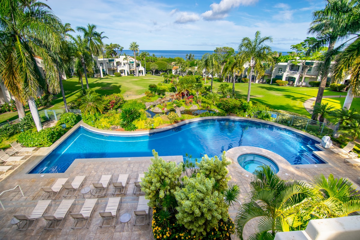 A tranquil outdoor space displays a large swimming pool adjacent to a hot tub, surrounded by palm trees and lush landscaping. Lounge chairs are arranged neatly on the pool deck, complementing the serene view of the manicured grounds and distant ocean.