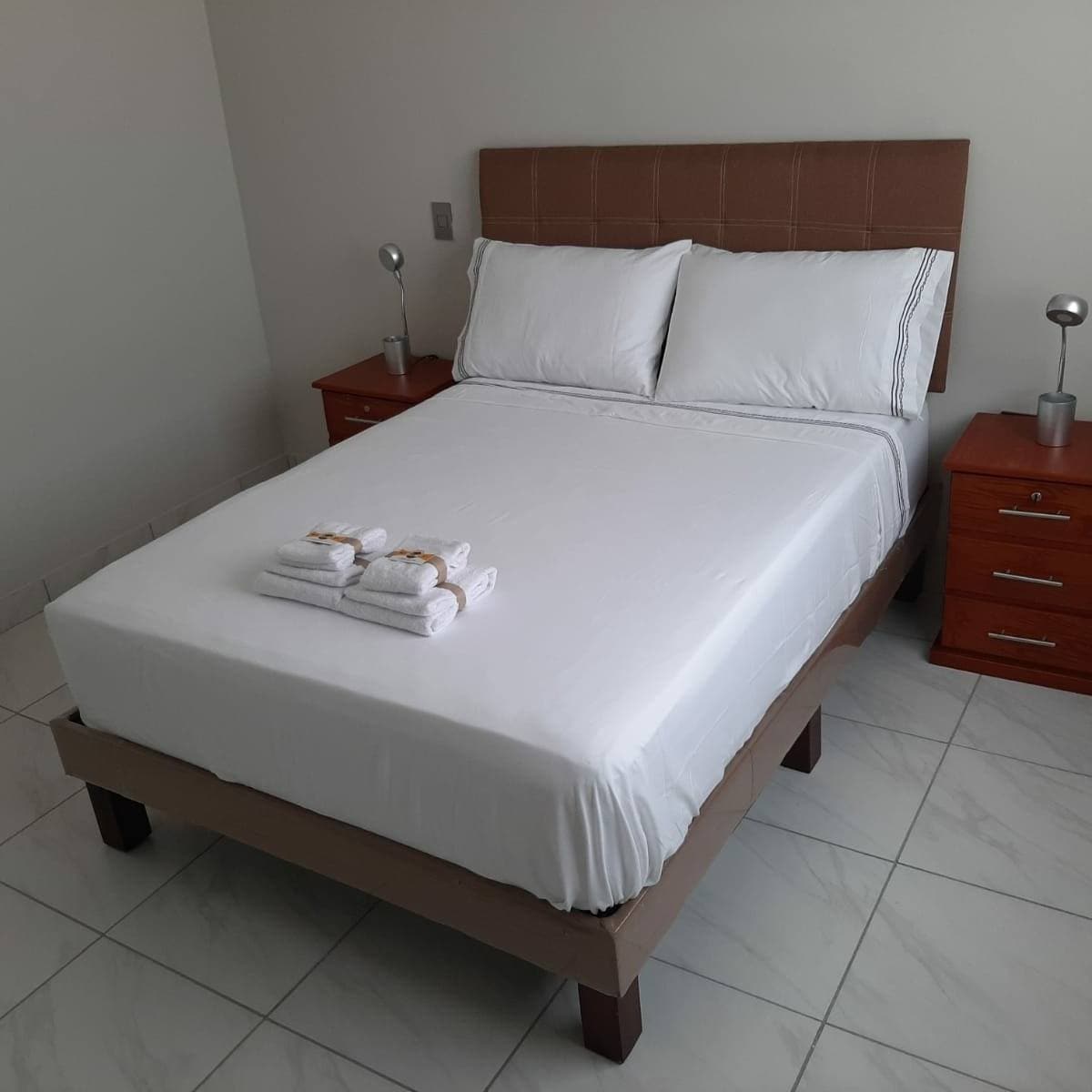 A spacious sleeping area features a comfortable bed with crisp linens, flanked by two wooden nightstands. Each nightstand is equipped with a sleek lamp. Neatly arranged towels are placed on the bed, enhancing the room's welcoming ambiance.
