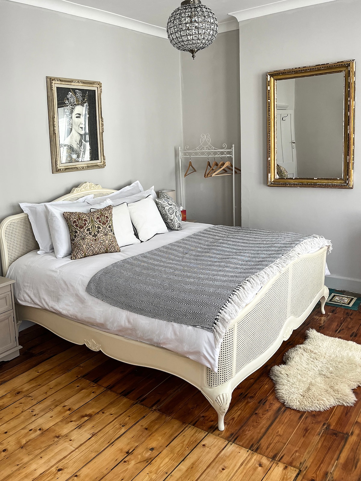 A spacious double bedroom features a superking bed, draped with a soft gray throw. A stylish mirror reflects light and enhances the room's airy feel. Stripped pine floorboards add warmth, while a hanging rack is visible in the background, providing practical storage.