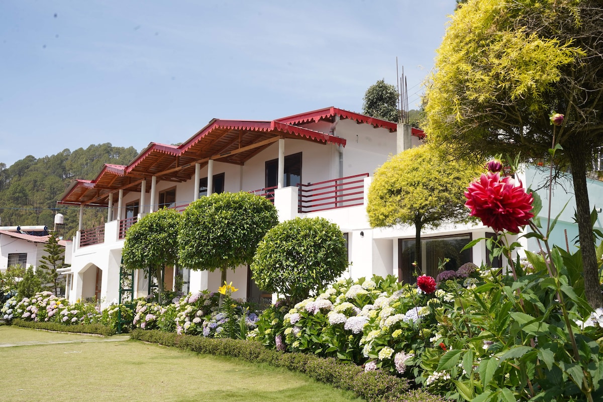 A well-maintained property is surrounded by lush greenery and vibrant flower beds. The two-story building features a red-roofed design with spacious balconies. Neatly trimmed hedges and colorful hydrangeas are visible in the garden, adding to the tranquil atmosphere of the area.