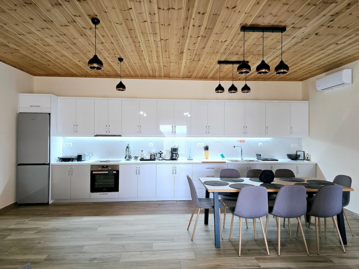 A modern kitchen is presented with a sleek design featuring white cabinetry and a wooden ceiling. Stainless steel appliances, including a refrigerator and oven, are complemented by a dining table surrounded by six chairs, creating a welcoming space for meal preparation and dining.