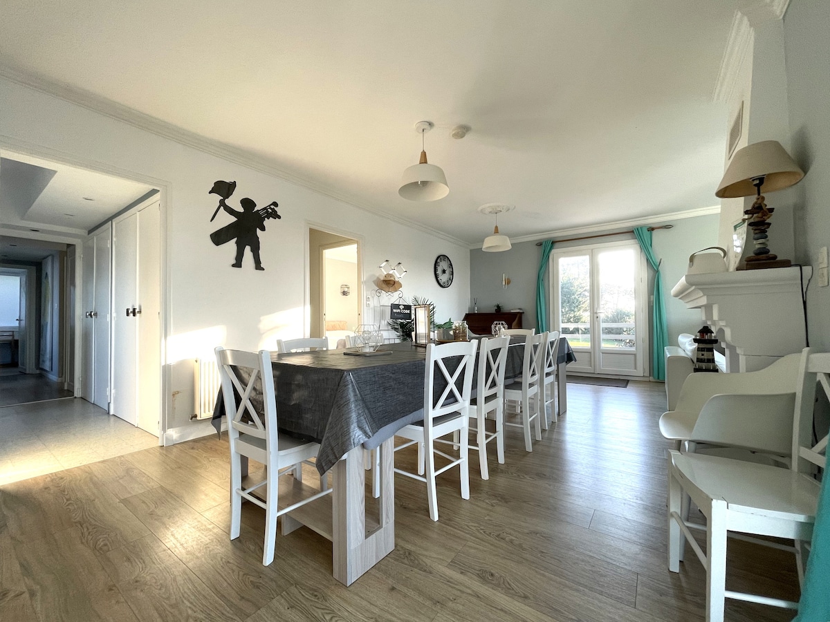 A spacious dining area features a long table surrounded by white chairs, with natural light streaming through large windows. A decorative figure on the wall adds character, while a soft color palette contributes to the inviting environment.