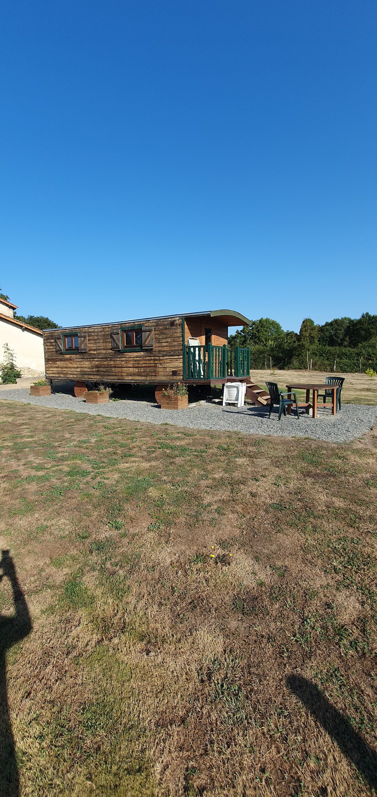 A wooden caravan is set in a tranquil outdoor space, with a gravel area featuring a dining table and chairs. The caravan's porch is accessible, inviting relaxation. Surrounding greenery adds to the peaceful atmosphere, and clear blue skies contribute to the serenity of the environment.