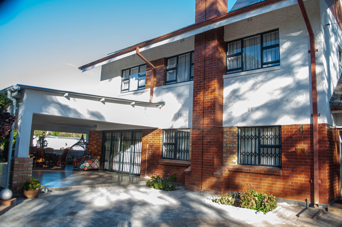 The exterior of the house features a two-story structure with brick and white walls. Large windows allow natural light to fill the interior. A covered patio provides a welcoming entrance, with a glimpse of the lush garden and colorful seating area visible in the background.