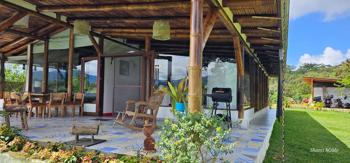 An expansive porch area features bamboo furniture and a lounge chair, complemented by a well-maintained garden. The open design allows for unobstructed views of the surrounding nature. A grill is positioned on one side, inviting outdoor dining experiences.