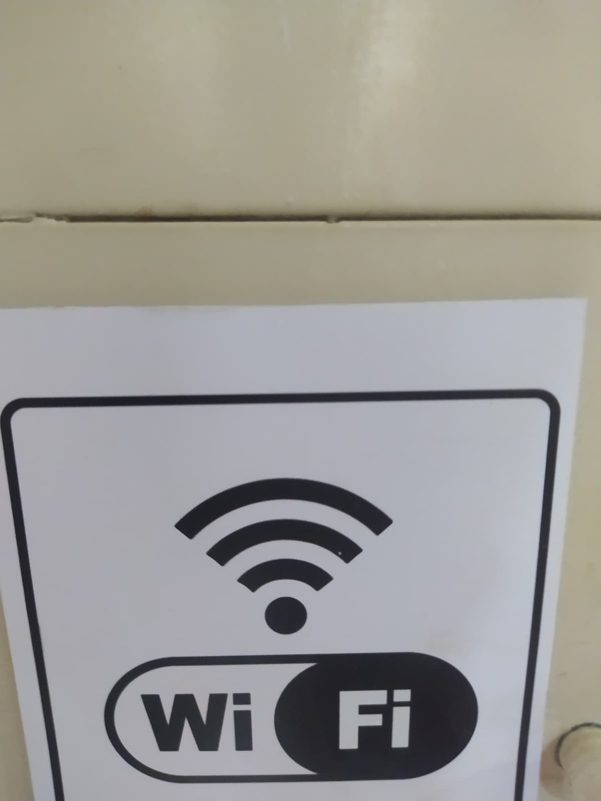 A white sign is displayed on a surface, featuring a black Wi-Fi symbol above the word 'Wi-Fi.' The design is simple and clear, indicating the availability of wireless internet in the space.